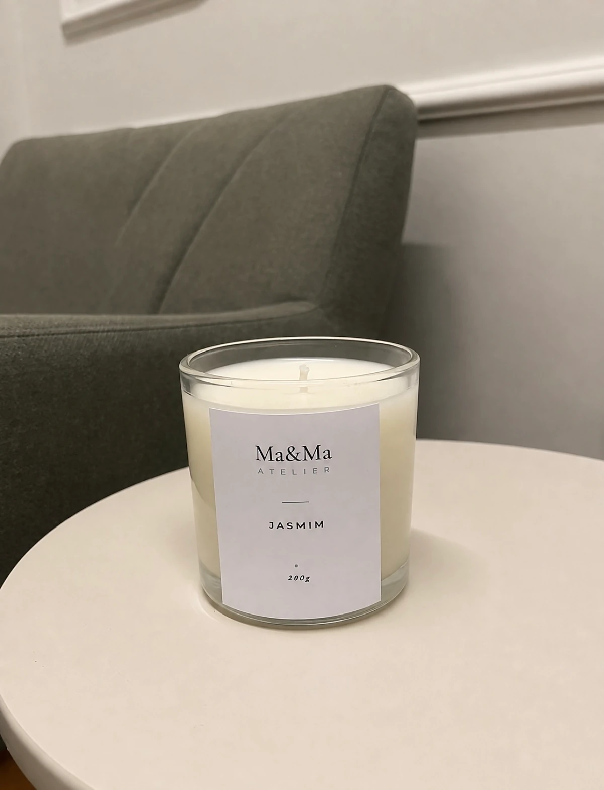 Vela Presença de Jasmim sobre mesa lateral branca, com uma poltrona verde ao fundo